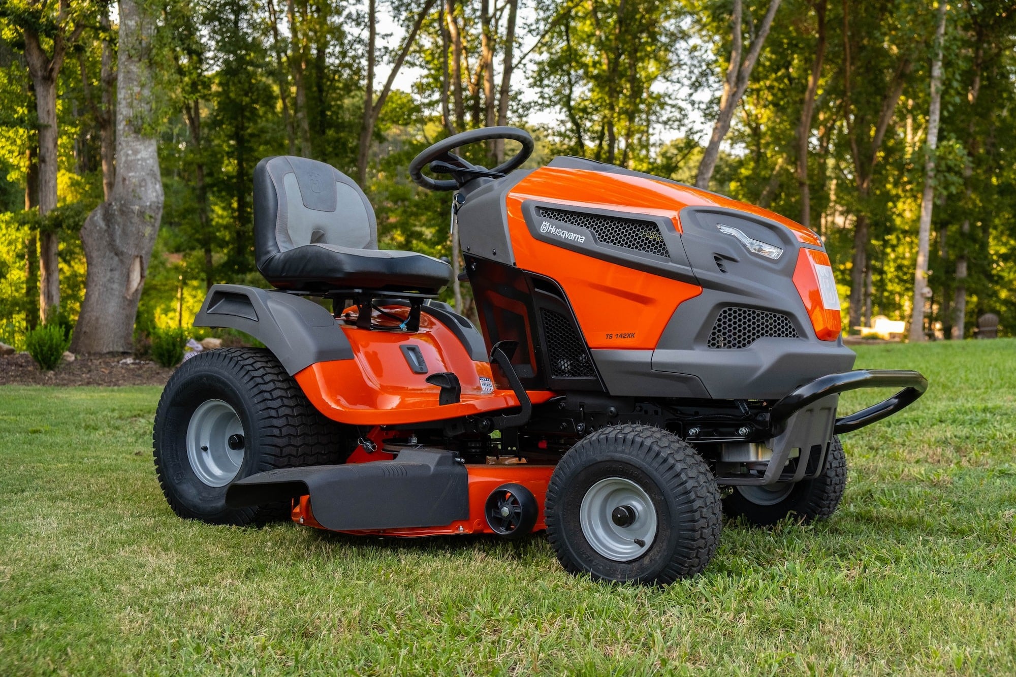 Red and black home use riding mower sitting on bright green grass, surrounded by green trees.