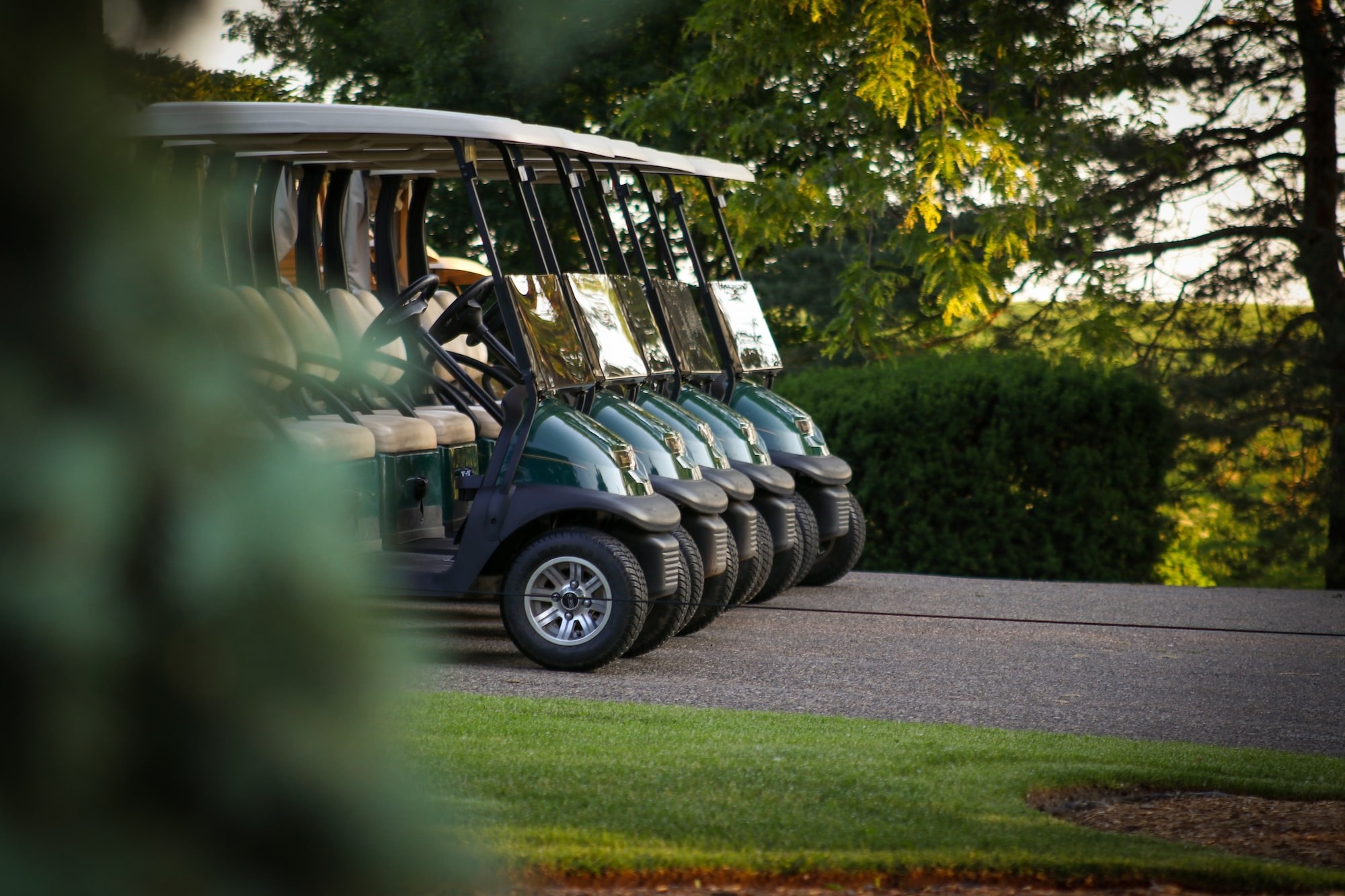 Line of five green two seater golf carts on a cart path surrounded by bueatiful greenery. 