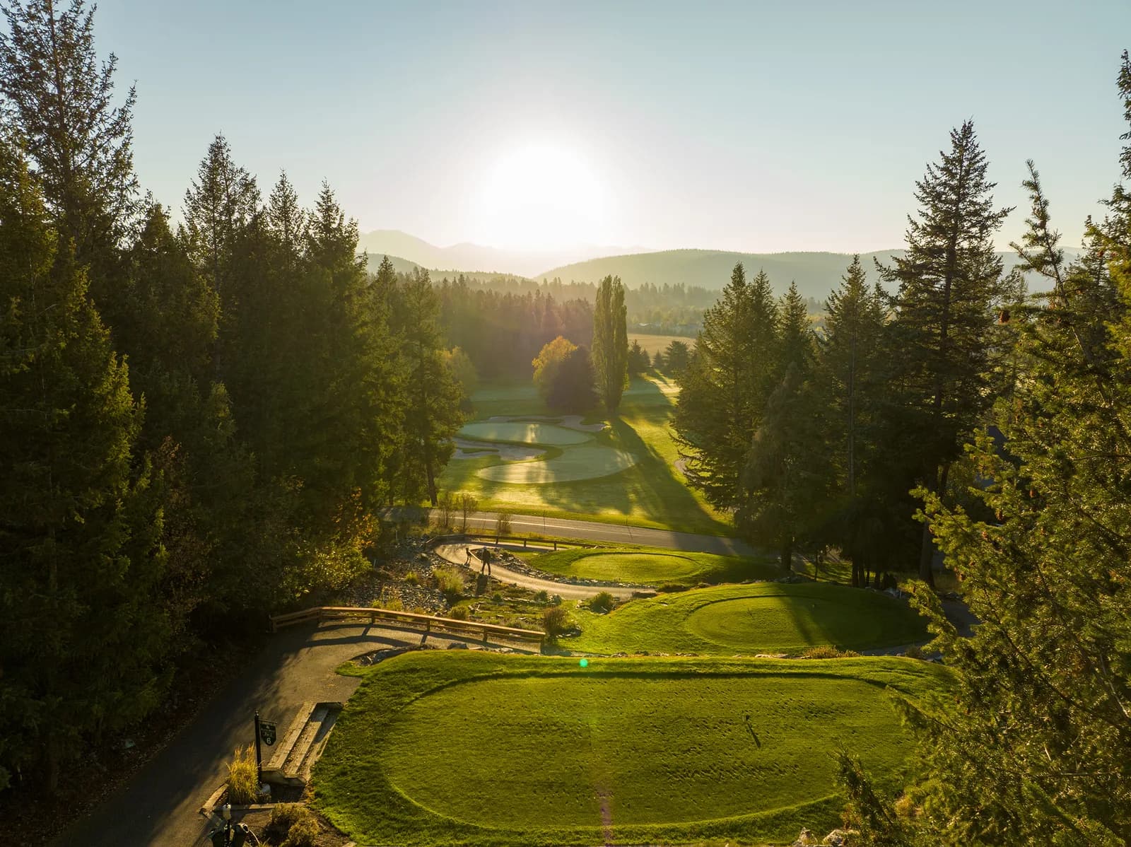 Looking down a northwest Montana hole at sunrise, tee boxes in the foreground, a pond and fairway below, forested hills beyond.