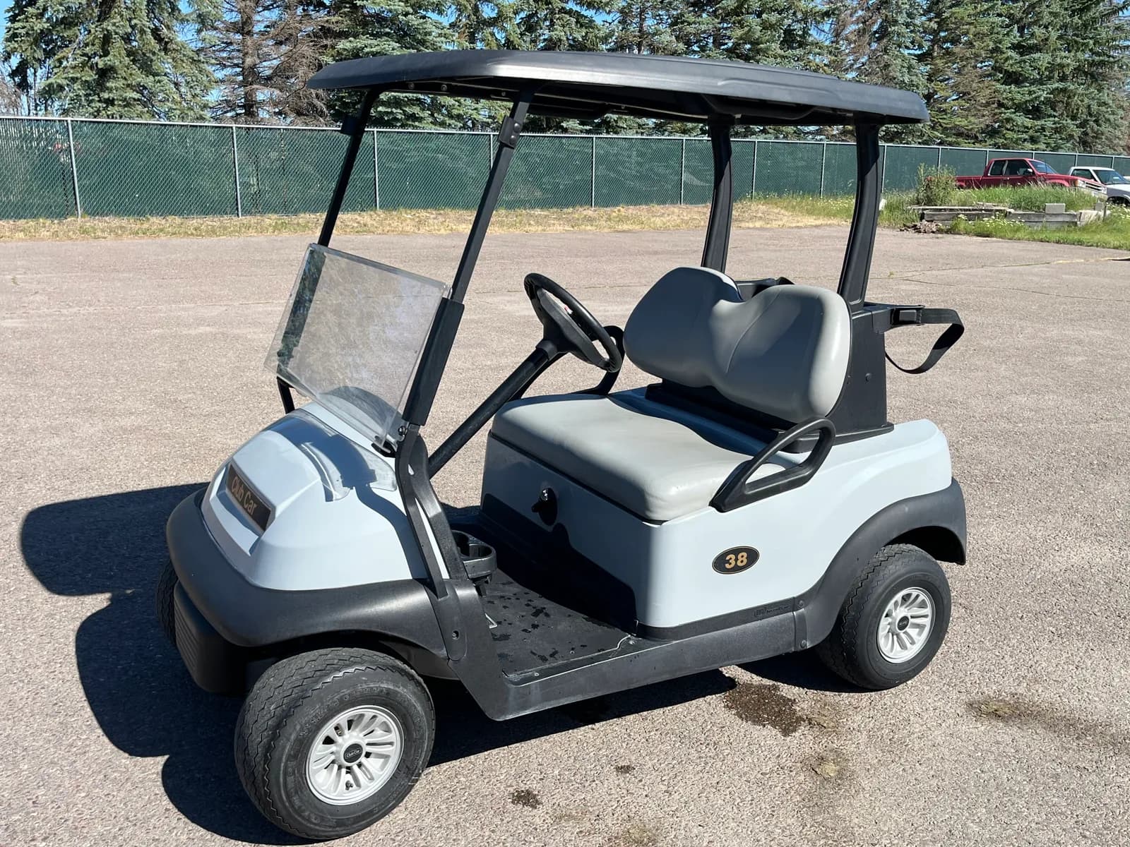 A white two seater Club Car rental cart, number 38, parked on asphalt in front of a chain link fence and pines.