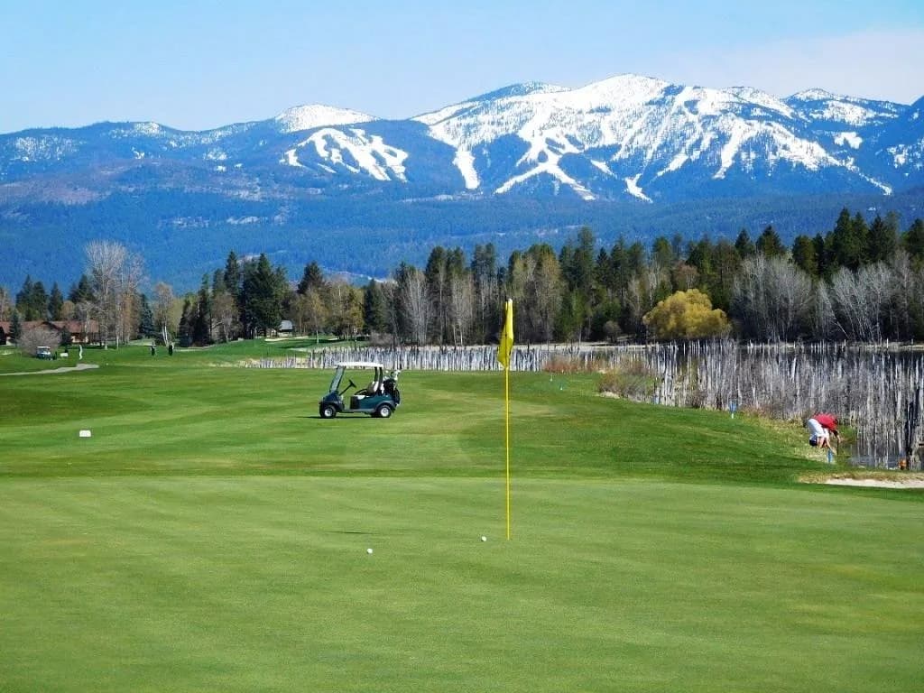 A putting green in early spring with a yellow flagstick, a cart parked on the fairway behind it, and a ski hill still under snow in the distance.