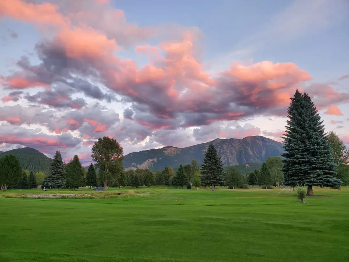 A Flathead Valley fairway at dusk, pink and grey clouds overhead and mountains behind the treeline.